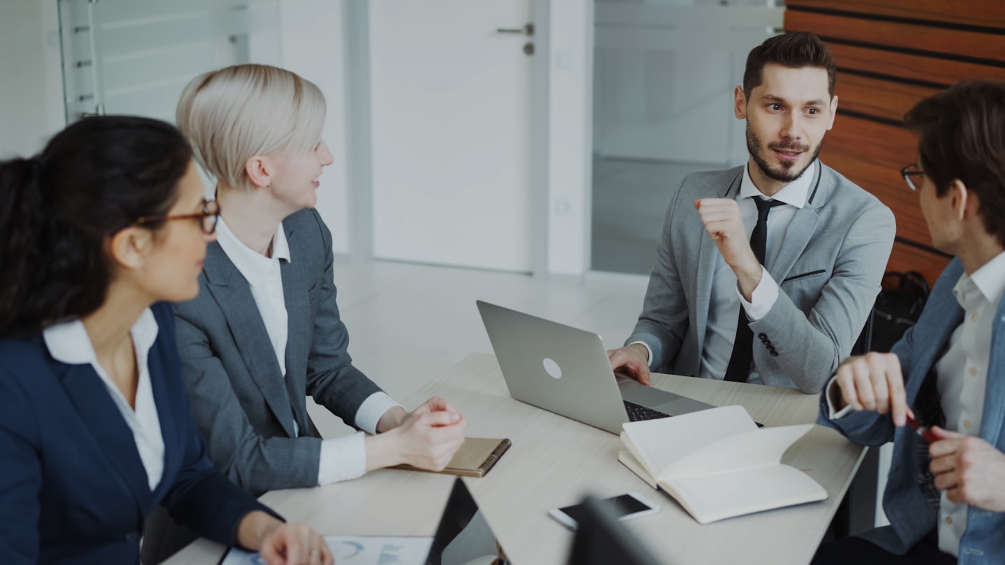 Professionals discuss business around a conference table with laptops and documents.